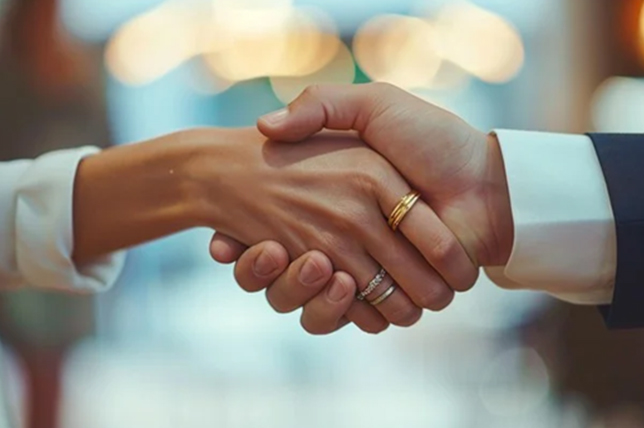A close-up of a handshake between two people, one wearing a suit. The background is softly blurred with warm lighting, conveying a sense of agreement and professionalism.